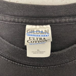 May include: Close-up of a dark gray t-shirt with a white tag. The tag reads "GILDAN ACTIVEWEAR ULTRA COTTON HEAVYWEIGHT" and "XL 100% COTTON PRESHRUNK MADE IN CANADA". The shirt is made of cotton.