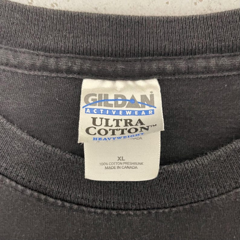 May include: Close-up of a dark gray t-shirt with a white tag. The tag reads "GILDAN ACTIVEWEAR ULTRA COTTON HEAVYWEIGHT" and "XL 100% COTTON PRESHRUNK MADE IN CANADA". The shirt is made of cotton.