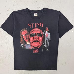 Op de afbeelding: Een zwart T-shirt met korte mouwen en een grafische print van de worstelaar Sting. Het ontwerp bevat de tekst STING in het rood, een groot rood masker en twee afbeeldingen van de worstelaar. Onder het masker staat een rood kader met de witte tekst nWo.