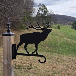 May include: Black metal elk silhouette hanging bracket. The bracket is mounted on a wooden post with a decorative finial. The elk is depicted with large antlers and is facing right. The text "PeCo Metal Decor" is visible on the bracket.