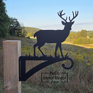 May include: Black metal deer silhouette mounted on a wooden post. The deer is standing on a decorative bracket with a curved design. The bracket is attached to a wooden post. The text "PeCo Metal Decor & more" is visible.