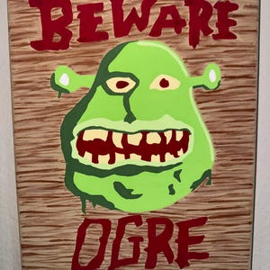 May include: A painted canvas sign featuring a green ogre face with a wide, toothy grin. The words "BEWARE OGRE" are written in red block letters above and below the ogre's face. The background has a wood grain texture.