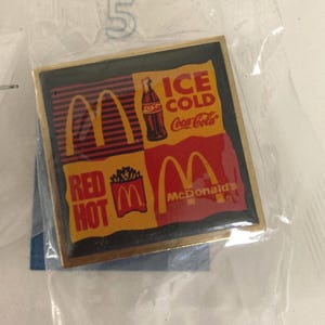 Puede incluir: Un pin cuadrado con el logotipo de McDonald's y la marca Coca-Cola. El pin tiene un borde dorado y un dise&ntilde;o con las palabras "ICE COLD Coca-Cola" y "RED HOT McDonald's". El pin est&aacute; en una bolsa de pl&aacute;stico transparente.