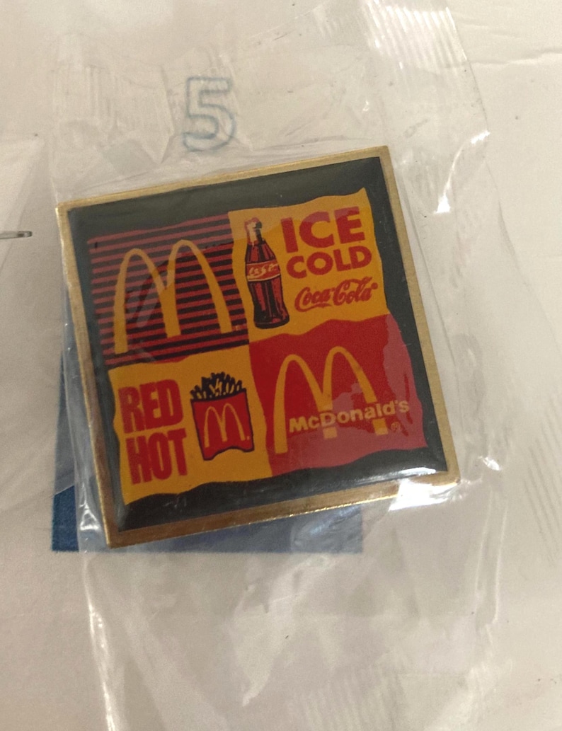 Puede incluir: Un pin cuadrado con el logotipo de McDonald's y la marca Coca-Cola. El pin tiene un borde dorado y un dise&ntilde;o con las palabras "ICE COLD Coca-Cola" y "RED HOT McDonald's". El pin est&aacute; en una bolsa de pl&aacute;stico transparente.