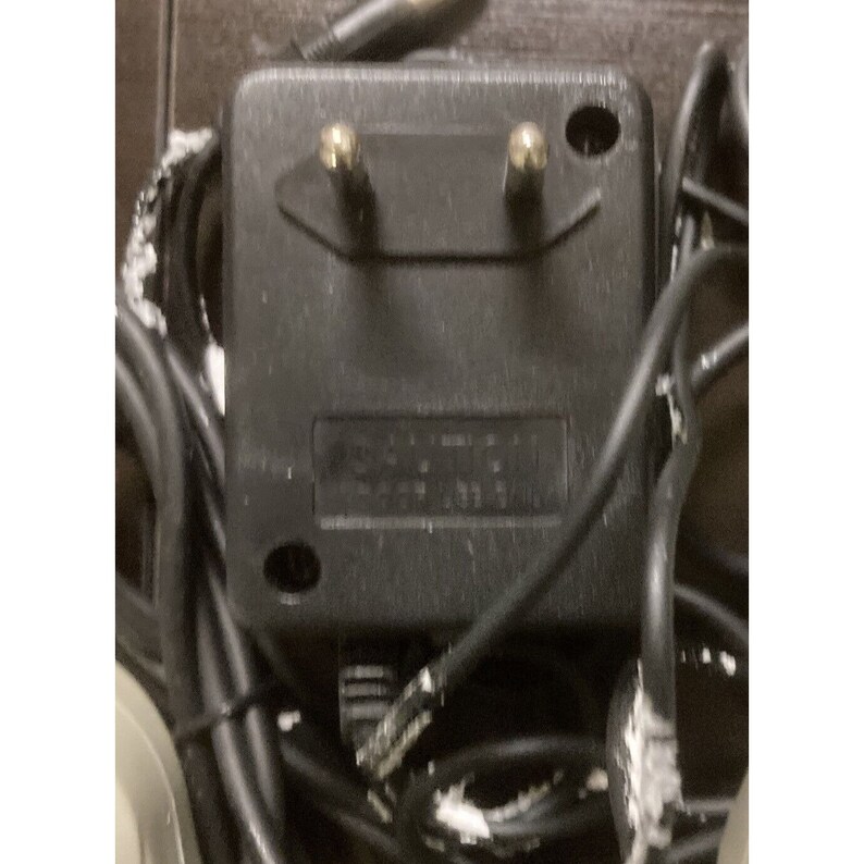 May include: A black power adapter with two gold-colored prongs. The adapter has a rectangular shape with a label and is surrounded by black cables. The image is a close-up shot of the adapter.