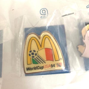 Puede incluir: Pin con el logotipo de los arcos dorados de McDonald's, un bal&oacute;n de f&uacute;tbol y un dise&ntilde;o de bandera. El pin es para la Copa del Mundo USA 94. El pin est&aacute; en una bolsa de pl&aacute;stico transparente.