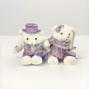 May include: Two white teddy bears dressed in formal attire. One bear wears a purple top hat, vest, and pants. The other bear wears a purple dress with lace trim and a bow. Both bears are holding hands.