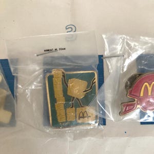 Puede incluir: Una colecci&oacute;n de pines promocionales de McDonald's, cada uno sellado en una bolsa de pl&aacute;stico transparente. Un pin presenta un personaje dorado sosteniendo bloques de letras, con la palabra "Scrabble" y el logotipo de McDonald's. Otro pin muestra un casco de f&uacute;tbol americano rojo con el logotipo de McDonald's.