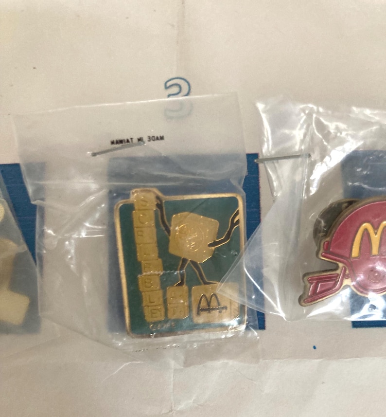 Puede incluir: Una colecci&oacute;n de pines promocionales de McDonald's, cada uno sellado en una bolsa de pl&aacute;stico transparente. Un pin presenta un personaje dorado sosteniendo bloques de letras, con la palabra "Scrabble" y el logotipo de McDonald's. Otro pin muestra un casco de f&uacute;tbol americano rojo con el logotipo de McDonald's.