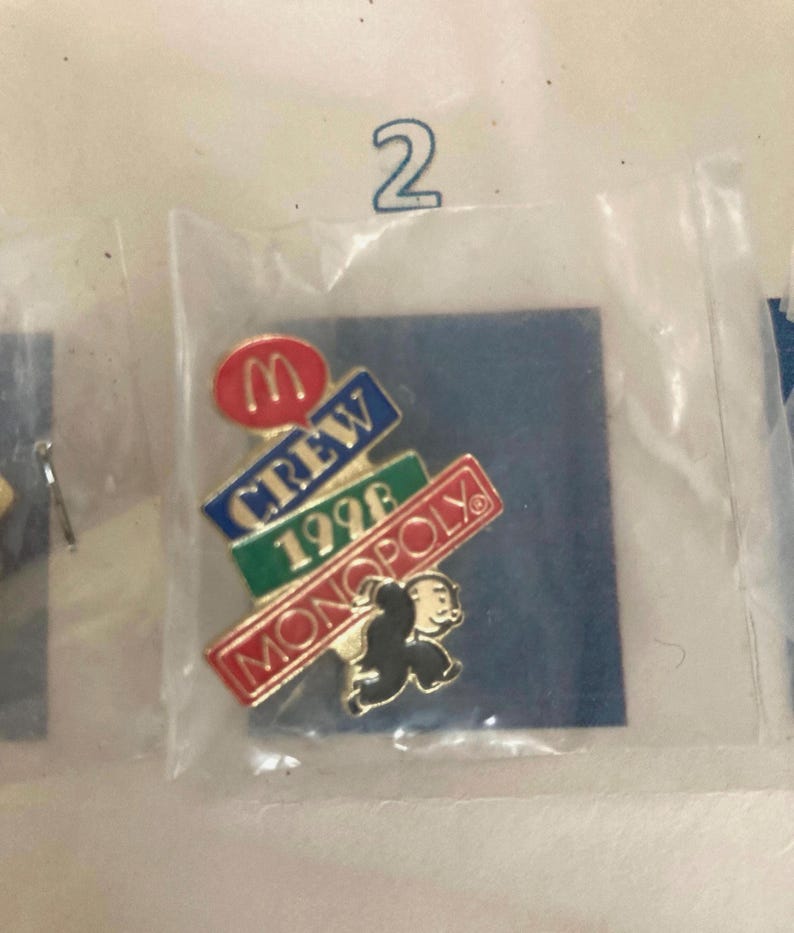 Puede incluir: Un pin con el logotipo de McDonald's y las palabras "CREW 1996 MONOPOLY" con un personaje del juego Monopoly. El pin est&aacute; en una bolsa de pl&aacute;stico sobre un fondo azul.
