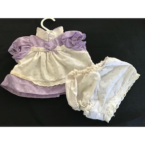 May include: A vintage doll dress and bloomers set. The dress features a white bodice with a small floral pattern and a lavender skirt and sleeves. The bloomers are white with a matching floral pattern and lace trim. The set is displayed on a white hanger.