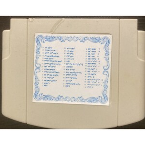 May include: A light gray rectangular cartridge with a white label featuring blue text and a decorative border. The label appears to list various items. The cartridge has a slightly textured surface.