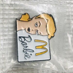 Puede incluir: Un pin tem&aacute;tico de Barbie con un personaje rubio y el logotipo de los arcos dorados de McDonald's. El pin tiene un fondo blanco con la palabra "Barbie" en escritura negra.