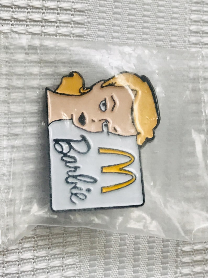 Puede incluir: Un pin tem&aacute;tico de Barbie con un personaje rubio y el logotipo de los arcos dorados de McDonald's. El pin tiene un fondo blanco con la palabra "Barbie" en escritura negra.