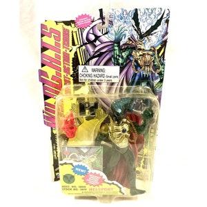 May include: Action figure of Helskont, a green and gold character with a red cape, packaged in a clear plastic bubble. The packaging features the text "S.P.A.W.N. - SUPER-ACTION-TERMS" and a warning about choking hazards.