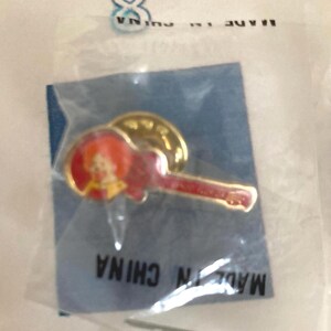 Puede incluir: Pin con forma de guitarra roja y detalles dorados, empaquetado en una bolsa de pl&aacute;stico transparente. El pin presenta una peque&ntilde;a imagen de una persona. El embalaje dice "MADE IN CHINA".