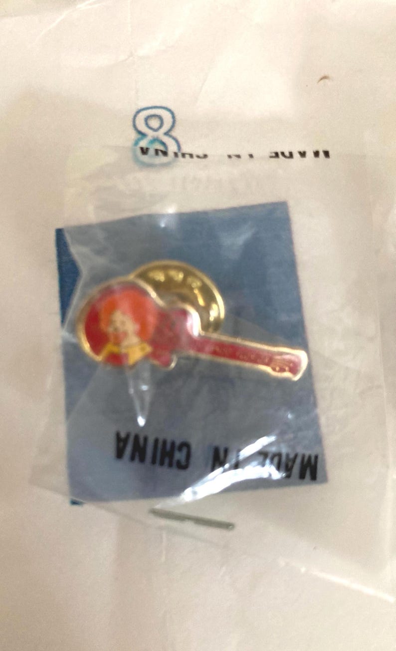 Puede incluir: Pin con forma de guitarra roja y detalles dorados, empaquetado en una bolsa de pl&aacute;stico transparente. El pin presenta una peque&ntilde;a imagen de una persona. El embalaje dice "MADE IN CHINA".