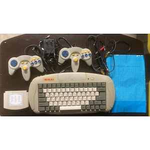 May include: A vintage NIKAI video game console set. The set includes a gray keyboard, two gray controllers with blue and yellow buttons, a game cartridge, and a blue grid-patterned pad. Cables and a power adapter are also included.