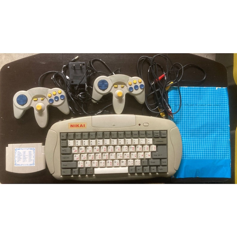 May include: A vintage NIKAI video game console set. The set includes a gray keyboard, two gray controllers with blue and yellow buttons, a game cartridge, and a blue grid-patterned pad. Cables and a power adapter are also included.