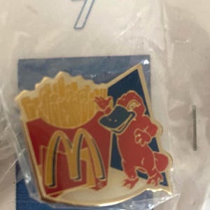Puede incluir: Pin con un personaje de ornitorrinco rojo con pico negro y un recipiente de patatas fritas doradas con el logotipo de McDonald's. El pin est&aacute; sobre un fondo azul.