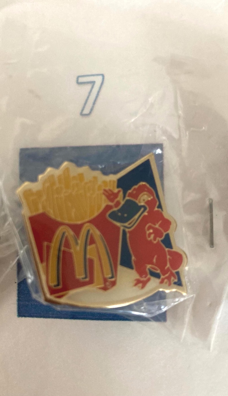 Puede incluir: Pin con un personaje de ornitorrinco rojo con pico negro y un recipiente de patatas fritas doradas con el logotipo de McDonald's. El pin est&aacute; sobre un fondo azul.