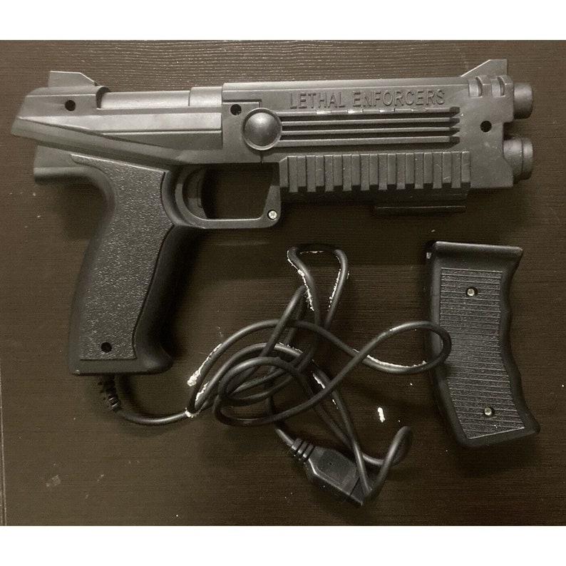 May include: Black plastic video game gun controller with the text "LETHAL ENFORCERS" on the side. Includes a separate grip and a wired connection. The controller has a textured grip and a trigger.
