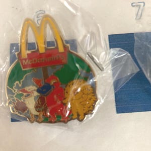 Puede incluir: Un colorido pin de McDonald's con el logotipo de los arcos dorados y personajes de dibujos animados. El pin est&aacute; en una bolsa de pl&aacute;stico. El pin tiene un fondo verde con un mapa y el logotipo de McDonald's.