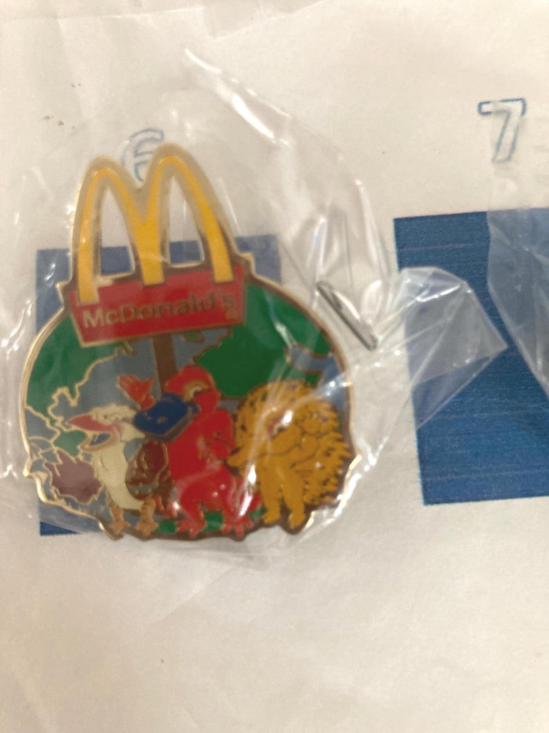 Puede incluir: Un colorido pin de McDonald's con el logotipo de los arcos dorados y personajes de dibujos animados. El pin est&aacute; en una bolsa de pl&aacute;stico. El pin tiene un fondo verde con un mapa y el logotipo de McDonald's.