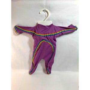 Könnte beinhalten: Ein lila Baby-Strampler mit Regenbogenbesatz an den Ärmeln, am Ausschnitt und am Frontreißverschluss. Der Strampler hängt an einem weißen Kleiderbügel vor einem weißen Hintergrund. Der Regenbogenbesatz umfasst Rot, Orange, Gelb, Grün, Blau und Lila.