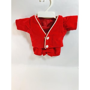 May include: A red velvet doll's outfit, comprising a short-sleeved jacket with white trim and two white buttons, and matching shorts. The outfit is suspended from a white plastic hanger. The outfit is likely for a doll.
