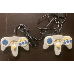 May include: Two gray video game controllers with blue and yellow buttons and a yellow joystick. Each controller has a black cord attached. The controllers are on a dark brown surface.