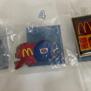 Puede incluir: Dos pines vintage de McDonald's con forma de casco de f&uacute;tbol. Un pin rojo con el logo "M" amarillo y el otro azul con un contorno blanco del estado. Ambos pines est&aacute;n en un paquete de pl&aacute;stico transparente.