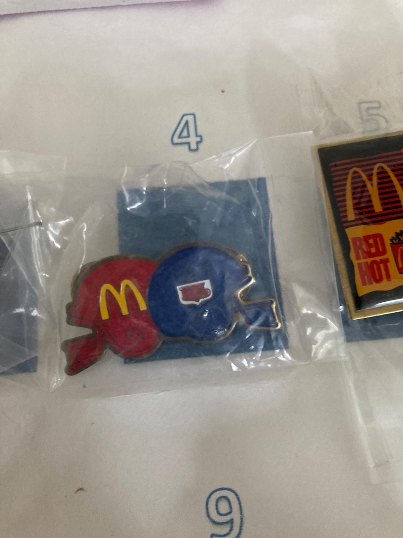 Puede incluir: Dos pines vintage de McDonald's con forma de casco de f&uacute;tbol. Un pin rojo con el logo "M" amarillo y el otro azul con un contorno blanco del estado. Ambos pines est&aacute;n en un paquete de pl&aacute;stico transparente.