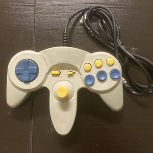 May include: A gray video game controller with a black cable. The controller features a blue directional pad, yellow and blue buttons, and a yellow joystick. The buttons are labeled with letters and numbers.