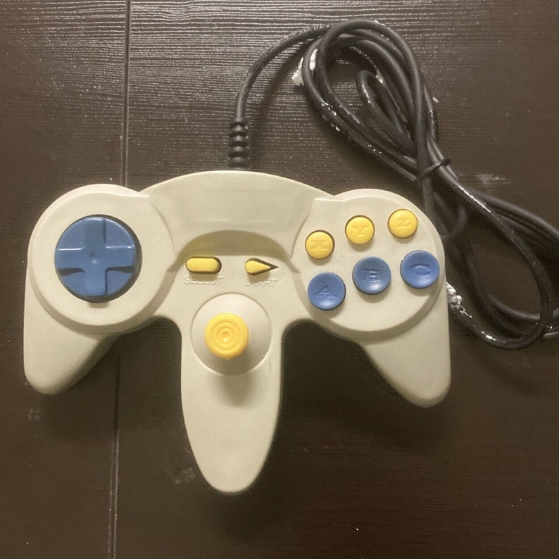 May include: A gray video game controller with a black cable. The controller features a blue directional pad, yellow and blue buttons, and a yellow joystick. The buttons are labeled with letters and numbers.