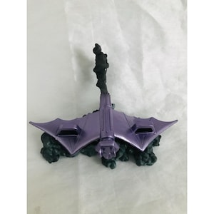 May include: A metallic purple bat-shaped vehicle with angular wings and a central fuselage, mounted on a dark, textured base that resembles a cloud of smoke or dark energy. The design features two rectangular intake vents on the wings.