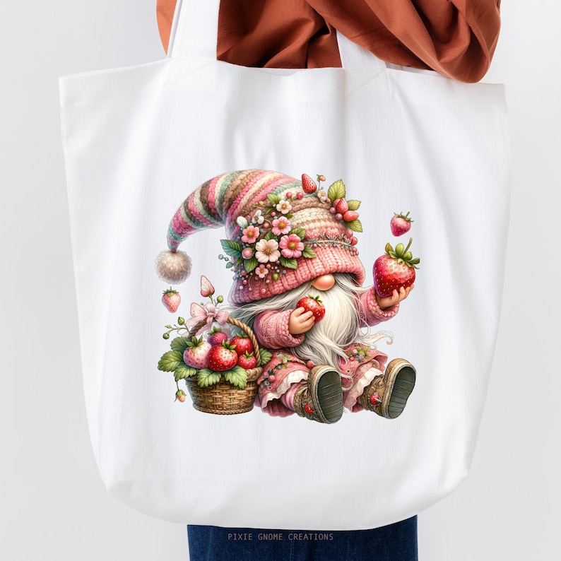 May include: White tote bag featuring a whimsical gnome design. The gnome, dressed in pink and holding strawberries, is seated near a basket of strawberries. The gnome wears a striped hat adorned with flowers. The text "PIXIE GNOME CREATIONS" is at the bottom.