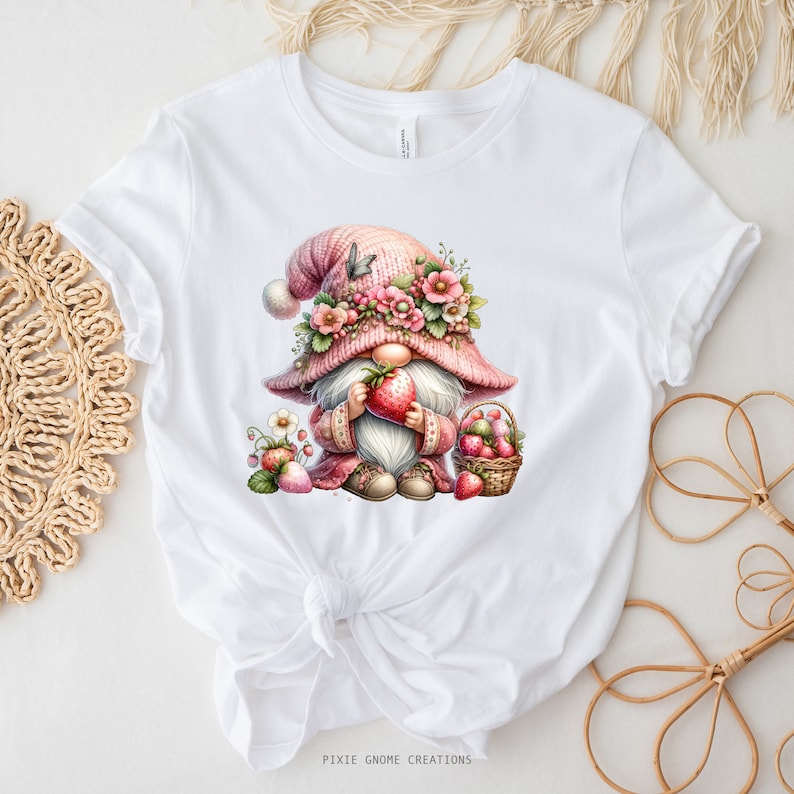 May include: White t-shirt featuring a watercolor illustration of a gnome holding a strawberry. The gnome wears a pink hat adorned with flowers and a matching outfit. A basket of strawberries is also shown. The text "PIXIE GNOME CREATIONS" is at the bottom.