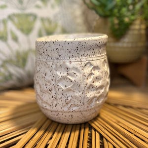 Könnte beinhalten: Weiße Keramiktasse mit gesprenkeltem Glase und einem erhabenen Prägemuster. Die Tasse steht auf einem geflochtenen Bambus-Tablett.