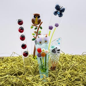 May include: Four vertical glass stakes featuring fused designs: one with red ladybugs, one with a brown owl on a branch, one with a light purple butterfly above colorful flowers, and one with a blue butterfly and a white patterned creature.