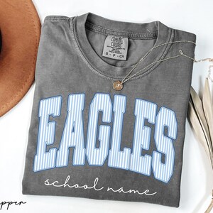 K&ouml;nnte beinhalten: Ein anthrazitfarbenes Kurzarm-T-Shirt mit dem Wort EAGLES in gro&szlig;en, hellblau-wei&szlig; vertikal gestreiften Blockbuchstaben. Unter dem Wort ist der Text school name in wei&szlig;er Schreibschrift gedruckt. Das T-Shirt hat einen Rundhalsausschnitt.