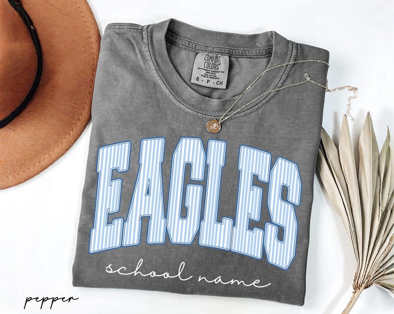 K&ouml;nnte beinhalten: Ein anthrazitfarbenes Kurzarm-T-Shirt mit dem Wort EAGLES in gro&szlig;en, hellblau-wei&szlig; vertikal gestreiften Blockbuchstaben. Unter dem Wort ist der Text school name in wei&szlig;er Schreibschrift gedruckt. Das T-Shirt hat einen Rundhalsausschnitt.