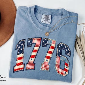 May include: Blue jean colored t-shirt with the number "1776" appliqued on the front in a patriotic design. The numbers feature red and white stripes, blue stars, and a red and white gingham pattern. A gold necklace rests on the shirt.