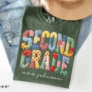 May include: A moss green crewneck t-shirt featuring the text SECOND GRADE in large, multi-colored, patterned fabric letters with a quilted appearance. Below the text, the name mrs. johnson is printed in a white, cursive, script-style font.