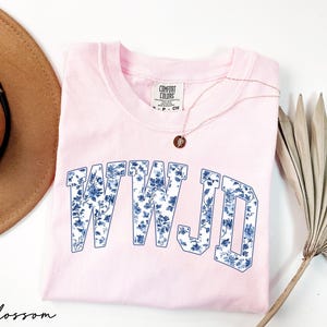 May include: A light pink crew neck sweatshirt with the letters "WWJD" in a blue floral pattern. A gold necklace with a small pendant rests on the shirt. A brown hat and a dried palm leaf are also in the image.