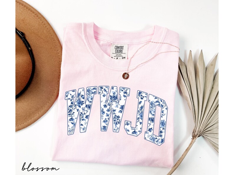 May include: A light pink crew neck sweatshirt with the letters "WWJD" in a blue floral pattern. A gold necklace with a small pendant rests on the shirt. A brown hat and a dried palm leaf are also in the image.