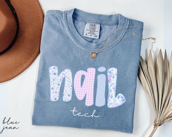 May include: A blue jean Comfort Colors t-shirt with the word "nail" in a floral, gingham, and striped pattern, and the word "tech" in a script font. A gold necklace with a pendant is on the shirt.