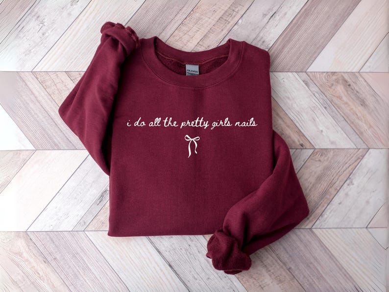 May include: A maroon sweatshirt with the text "i do all the pretty girls nails" in white script, with a bow graphic underneath. The sweatshirt is folded on a light wood-grain surface.