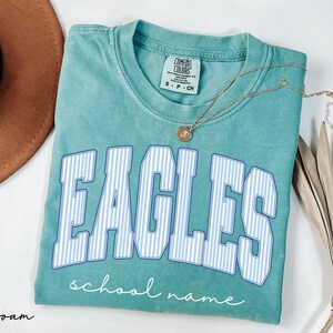 K&ouml;nnte beinhalten: Ein kurz&auml;rmeliges T-Shirt in Meerschaumgr&uuml;n mit Rundhalsausschnitt. Auf der Vorderseite steht das Wort EAGLES in gro&szlig;en, wei&szlig;-blau gestreiften Blockbuchstaben mit blauem Umriss und darunter der Text school name in wei&szlig;er Schreibschrift.