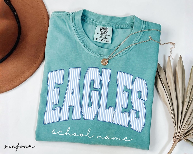 K&ouml;nnte beinhalten: Ein kurz&auml;rmeliges T-Shirt in Meerschaumgr&uuml;n mit Rundhalsausschnitt. Auf der Vorderseite steht das Wort EAGLES in gro&szlig;en, wei&szlig;-blau gestreiften Blockbuchstaben mit blauem Umriss und darunter der Text school name in wei&szlig;er Schreibschrift.